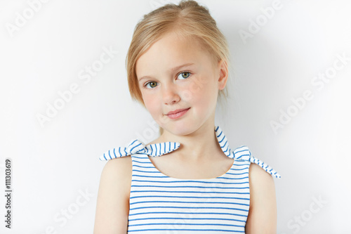 Canvas Print Close up shot of beautiful blonde Caucasian little girl wearing striped dress, looking at the camera with adorable smile, posing against white copy space concrete wall