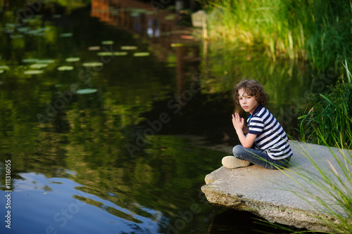 Fototapeta Naklejka Na Ścianę i Meble -  Pensive little boy sitting on the edge of the pond.