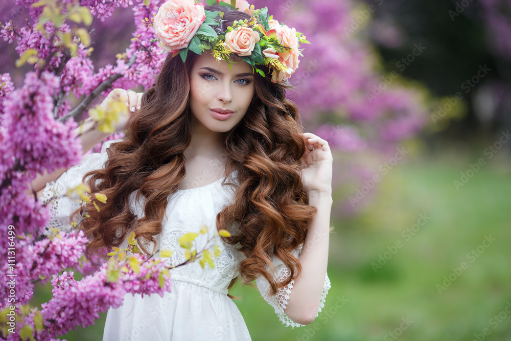 Naklejka premium Spring portrait of a beautiful woman in a wreath of flowers,long curly hair red color,gray eyes,beautiful smile,wearing a white summer dress,posing near a bush blooming pink flowers outdoors in spring
