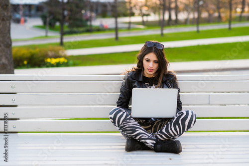 Portrait of a happy female student sitting on the bench with laptop outdoors and looking at camera