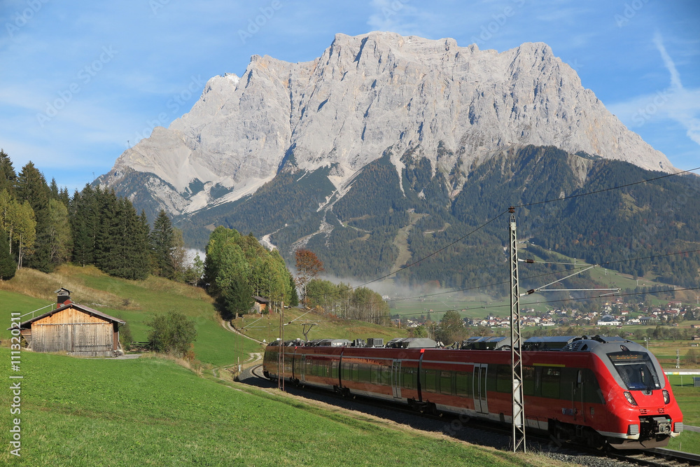 View of a train traveling through green fields with Mountain Zugspitze in the background on a beautiful sunny day in Lermoos, Tirol Austria ~ Magnificent summer scenery of idyllic Tyrolean countryside