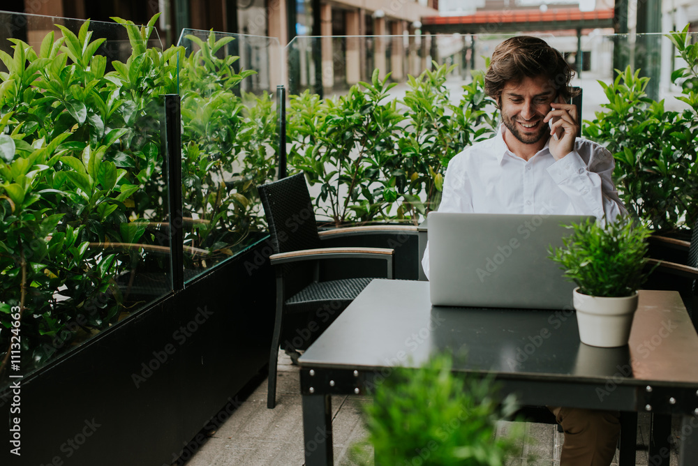 Smiling businessman in outdoor cafe working on laptop and talking on phone Smiling businessman in outdoor cafe working on laptop and talking on phone