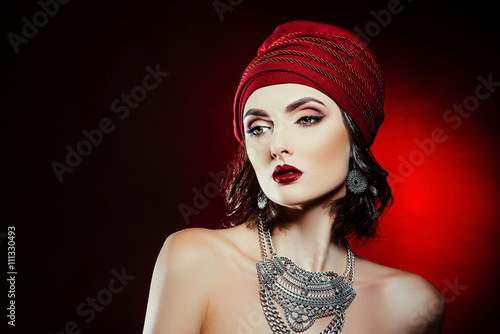 Beautiful brunette girl with makeup and hair in the form of a fortuneteller. With hats and metal decoration on the neck posing in the studio on a red background