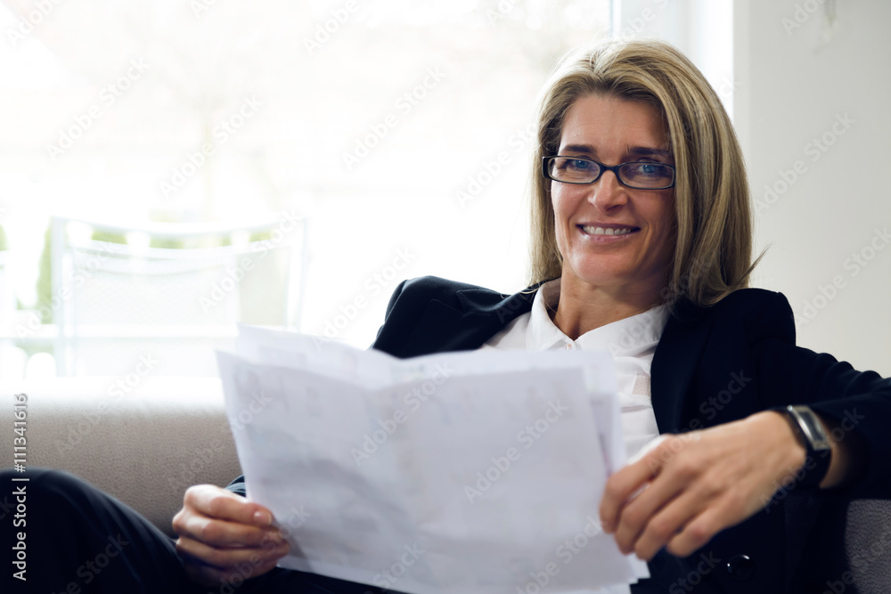 Portrait of attractive middle-aged business woman sitting on modern sofa at home and smiling into the camera. Confident woman with black suit reading white papers in bright environment.