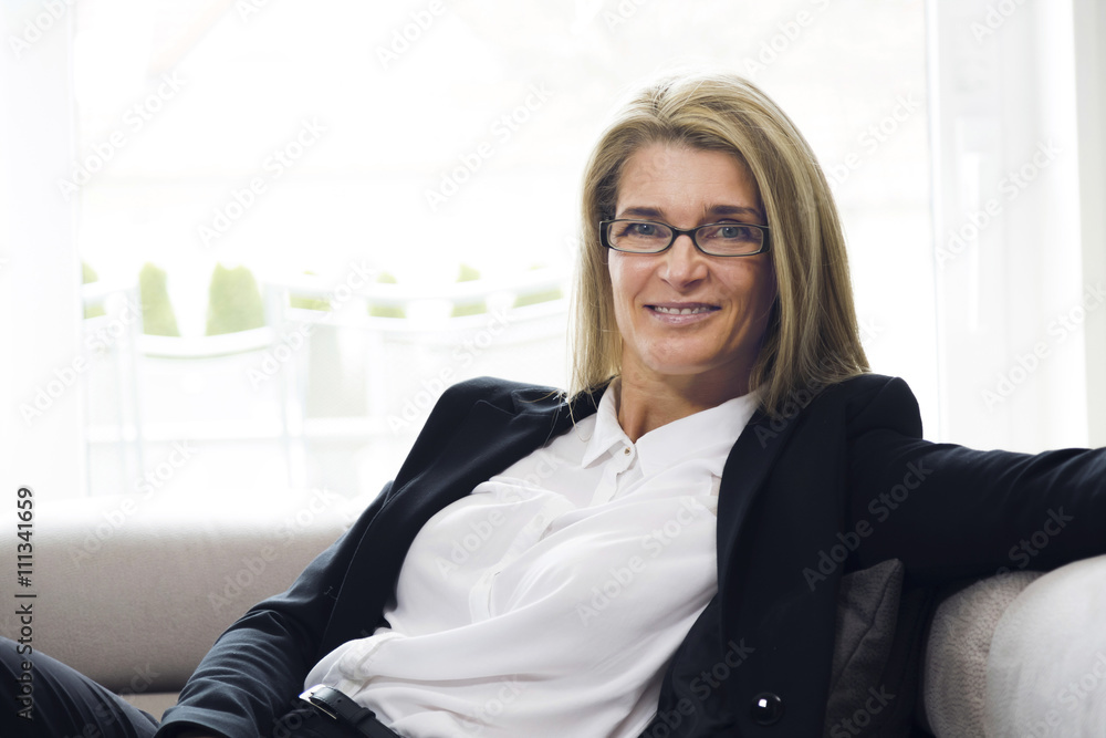 Portrait of attractive middle-aged business woman sitting on modern sofa at home and smiling into the camera. Confident woman with black suit in bright environment. Portrait of attractive middle-aged business woman sitting on modern sofa at home and smiling into the camera. Confident woman with black suit in bright environment.