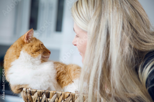 Cute cat with orange fur sitting in a round woven straw basket. Blonde attractive middle-aged woman cuddling with cat.