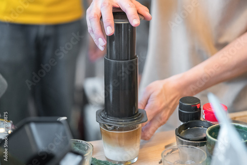 Step by step aero press coffee preparation Bearded barista in blue jeans shirt press aeropress to fill glass with beverage Professional coffee brewing cafe shop

 photo