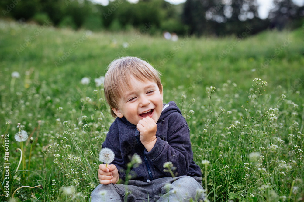 Happy smiling toddler boy playing in a blooming garden on a meadow green grass during summer day Happy smiling toddler boy playing in a blooming garden on a meadow green grass during summer day