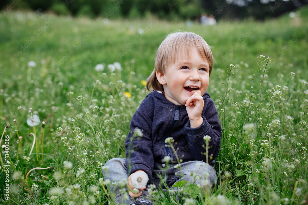Happy smiling toddler boy playing in a blooming garden on a meadow green grass during summer day Happy smiling toddler boy playing in a blooming garden on a meadow green grass during summer day