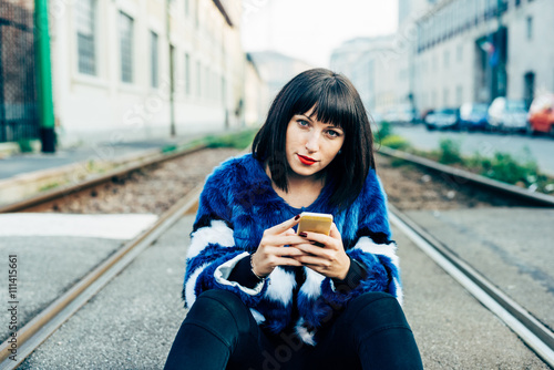 Young beautiful eastern black hair woman sitting on the floor, holding a smart phone, looking in camera pensive - technology, social network, communication concept