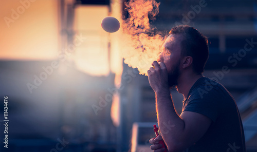 young man with beard on blurry background sunset sky, making soap bubbles with smoke inside with the aid of vape