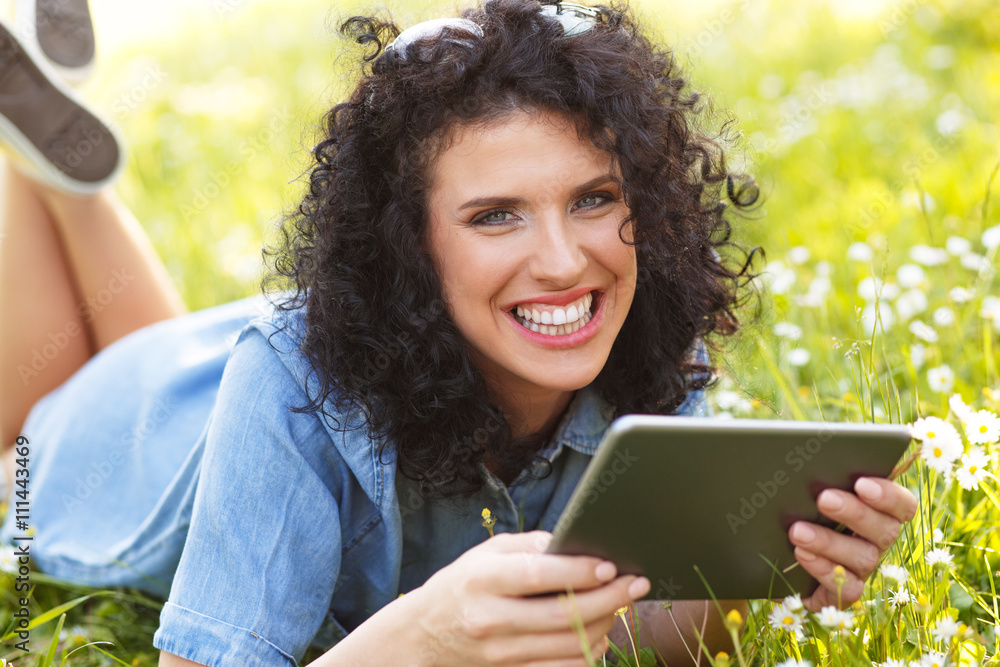 Pretty curly hair woman laying on grass with white flowers around her. She is using a tablet. Pretty curly hair woman laying on grass with white flowers around her. She is using a tablet.
