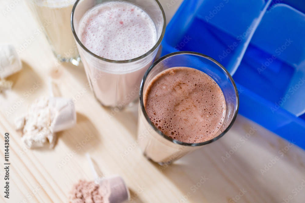 Three resh milk, chocolate, blueberry and banana drinks on table near protein shaker and meal box

