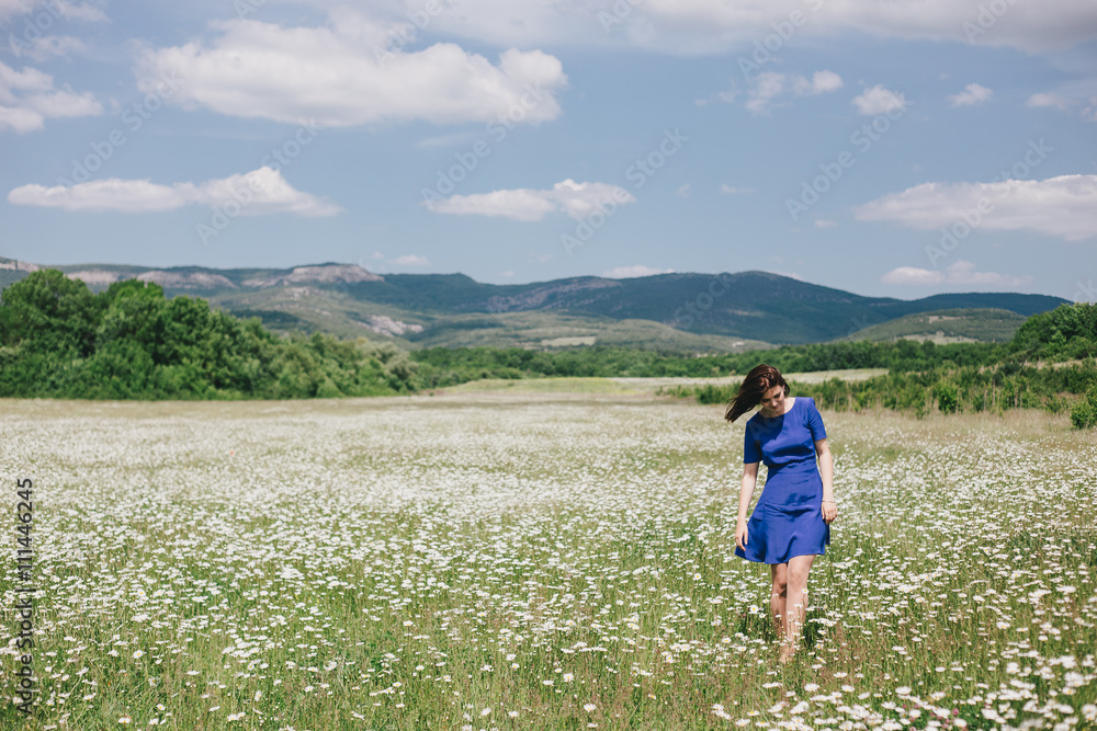 Young beautiful woman in a blue dress enjoying chamomile field among mountains. Summer mood