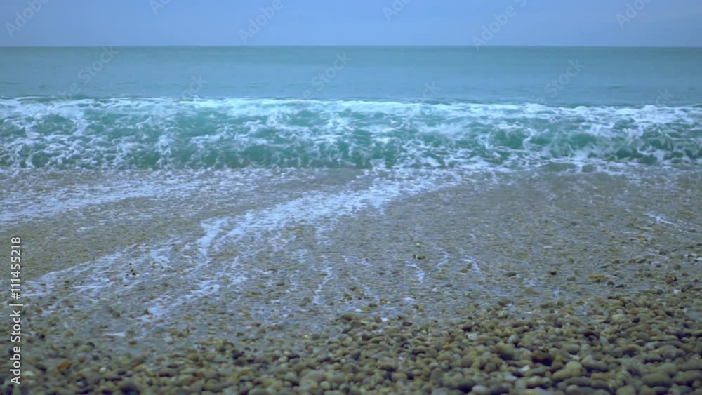 Foamy ocean wave rolling ashore in slowmotion, pebble beach under foggy sky Foamy ocean wave rolling ashore in slowmotion, pebble beach under foggy sky