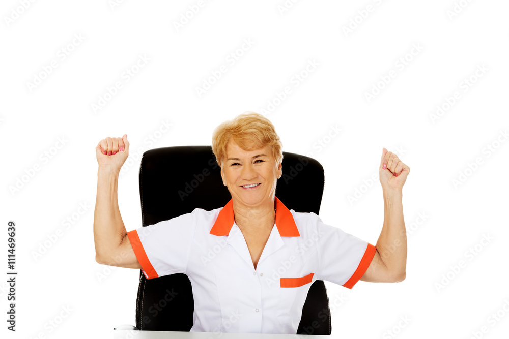 Happy elderly female doctor or nurse sitting behind the desk withd hands up Happy elderly female doctor or nurse sitting behind the desk withd hands up