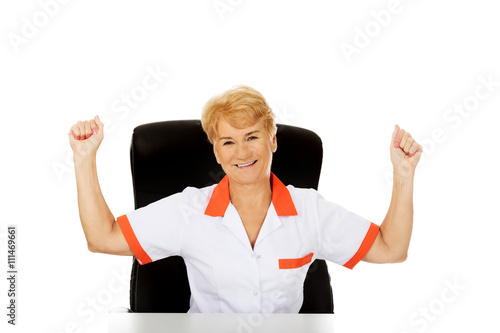 Happy elderly female doctor or nurse sitting behind the desk withd hands up photo