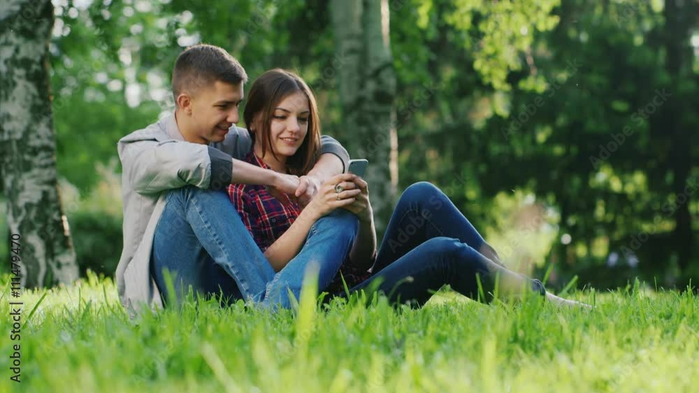 Man and woman hugging, sitting on the grass in the park. Looking at mobile phone screen. Communicate, smiling Man and woman hugging, sitting on the grass in the park. Looking at mobile phone screen. Communicate, smiling