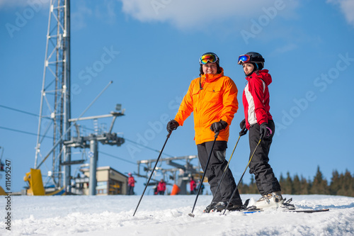 Couple holding skis standing together at a winter resort with ski lifts and blue sky in background. Man is wearing orange jacket, female in red jacket, both is wearing helmet and goggles.