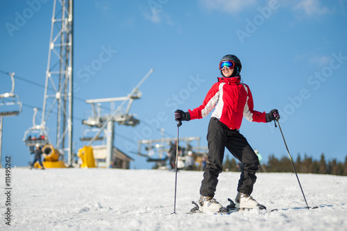 Woman wearing helmet, red jacket and ski goggles standing with skis on mountain top at a winter resort in sunny day with ski lifts and blue sky in background. Bukovel, Ukraine