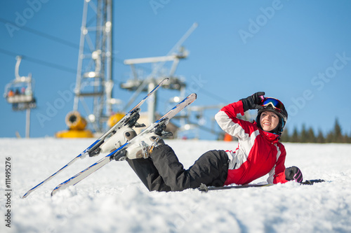 Female raising her ski glasses and looking away, lying with skis on snowy at mountain top in sunny day with ski lifts and blue sky in background.