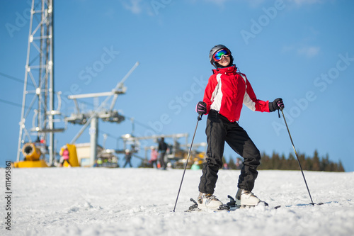 Young female skier wearing helmet, red jacket and ski goggles standing with skis on mountain top at a winter resort in sunny day with ski lifts and blue sky in background.