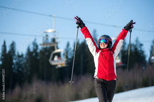 Happy female skier wearing helmet, red jacket and ski goggles standing on snowy slope with hands raised up in sunny day with forest and blue sky in background. Carpathian, Bukovel, Ukraine