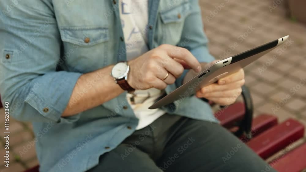 Pretty young hipster man sitting on a bench using tablet outdoors. Close up view of hands Pretty young hipster man sitting on a bench using tablet outdoors. Close up view of hands