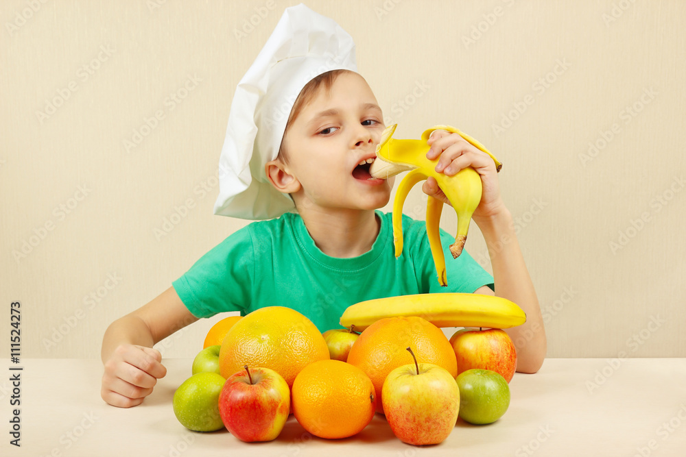 Little funny boy eat fresh banana at the table with fruits Little funny boy eat fresh banana at the table with fruits