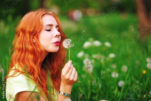 beautiful red-haired young woman blowing away dandelion outdoors