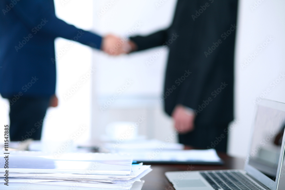 Documents and laptop on the table. Business people shaking hands on the background Documents and laptop on the table. Business people shaking hands on the background