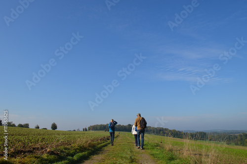 Hikers walking on trail through rural landscape with meadows and fields on rolling hills in Wallonia on sunny autumn day, Durnal, Yvoir