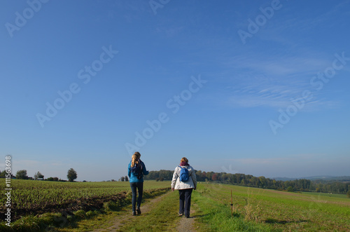 Woman and girl walking on trail through rural landscape with meadows and fields on rolling hills in Wallonia on sunny autumn day, Durnal, Yvoir