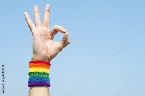 Hand of athlete giving OK sign with gay pride rainbow colors wristband against bright blue sky photo