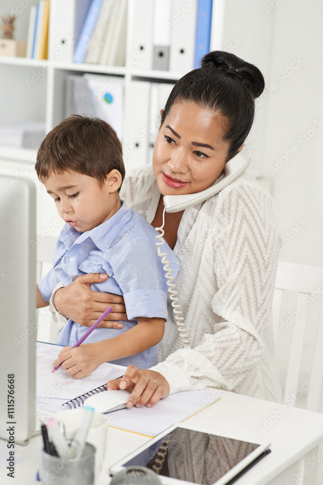 Indonesian business lady working in office with son on her knees Indonesian business lady working in office with son on her knees
