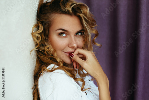 Beautiful blonde girl with long hair. With hair and makeup, posing in the morning in an apartment on the background of purple curtains in a white shirt. The light from the window, soft focus