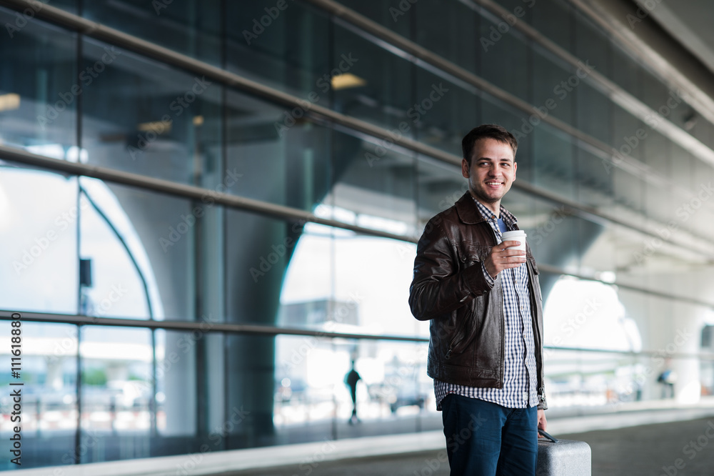 Stylish handsome young male traveller with bristle standing outdoors. Man wearing jacket and shirt. Smiling person looking to camera holding cup of coffee Stylish handsome young male traveller with bristle standing outdoors. Man wearing jacket and shirt. Smiling person looking to camera holding cup of coffee
