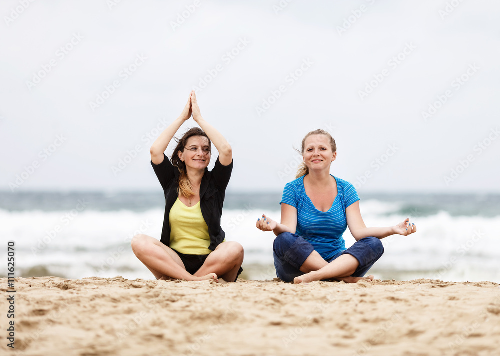 Two smiling young women sitting and relax in lotus pose outdoors. Two happy girls sitting outdoors. Selective focus on the models. Two smiling young women sitting and relax in lotus pose outdoors. Two happy girls sitting outdoors. Selective focus on the models.