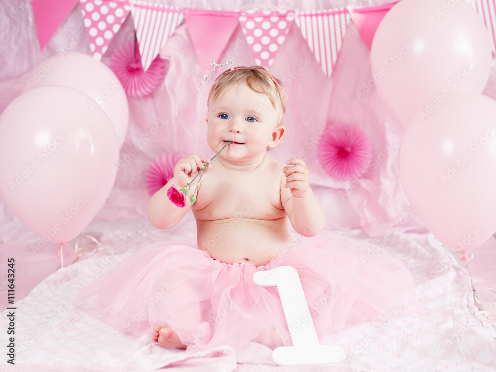 Fototapeta premium Portrait of cute adorable Caucasian baby girl with blue eyes in pink tutu skirt celebrating her first birthday with flower, gourmet cake and balloons looking away, cake smash first year concept