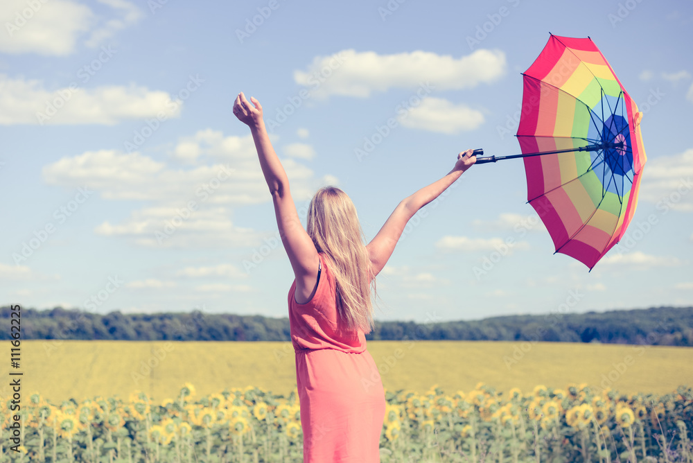 Back view of beautiful young lady holding multicolored umbrella in sunflower field and blue sky outdoors. - obrazy, fototapety, plakaty 