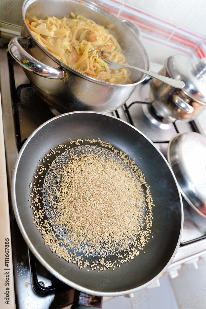 Tasty homemade sesame seeds in metal pan and noodles cooked with shrimps standing on gas stove Tasty homemade sesame seeds in metal pan and noodles cooked with shrimps standing on gas stove