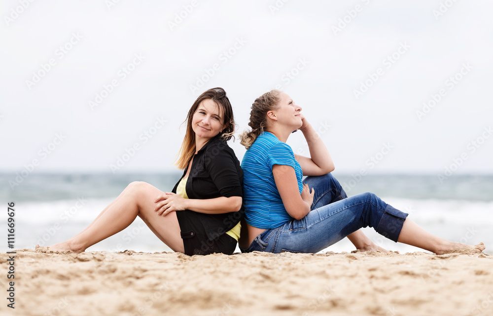 Two pretty young beautiful smiling women sitting with his back to each other and resting on the sand on the seashore. Two girls resting on the beach. Two girls outdoors. Selective focus on the models. Two pretty young beautiful smiling women sitting with his back to each other and resting on the sand on the seashore. Two girls resting on the beach. Two girls outdoors. Selective focus on the models.