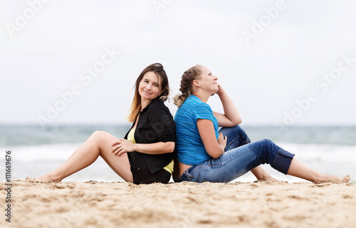 Two pretty young beautiful smiling women sitting with his back to each other and resting on the sand on the seashore. Two girls resting on the beach. Two girls outdoors. Selective focus on the models.