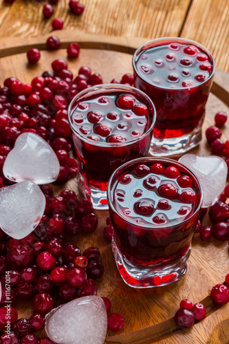 Cold cranberry drink in glasses, cranberries and pieces of ice on wooden background.