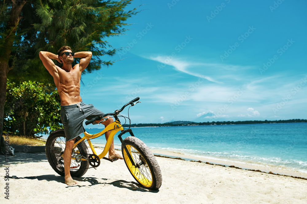 Fitness Male Model With Bike Sunbathing On Sun, Enjoying Summer Travel  Vacation. Handsome Sexy Man In Sunglasses With Muscular Body, Healthy Tan  Skin Tanning Near Bicycle On Sand Beach. Summertime foto de