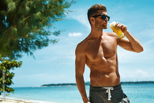 Healthy Drink. Handsome Fitness Male Model Having Fun, Enjoying Travel Vacation. Portrait Of Athletic Sexy Man With Muscular Body Drinking Refreshing Juice Cocktail On Tropical Sea Beach. Summertime