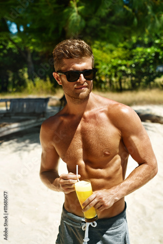 Summer Relax. Portrait Of Athletic Sexy Man With Muscular Body Drinking Fresh Juice Smoothie Cocktail On Tropical Beach. Handsome Fitness Male Model Sunbathing, Enjoying Refreshing Drink On Vacation