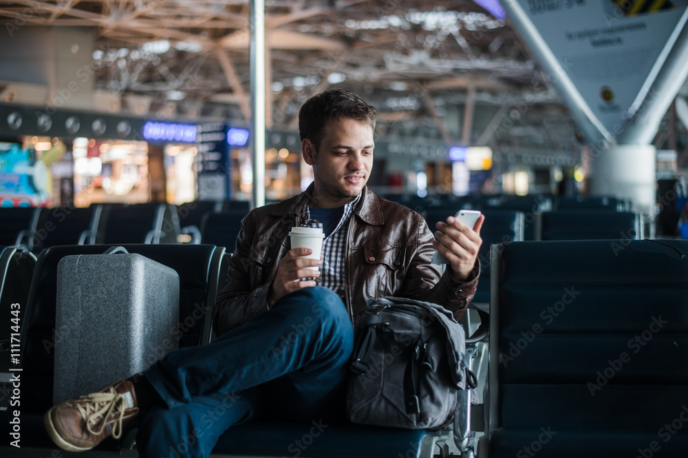 Handsome smiling man in casual wear holding luggage and messaging through his mobile phone while sitting at the hall of airport with coffee cup Handsome smiling man in casual wear holding luggage and messaging through his mobile phone while sitting at the hall of airport with coffee cup