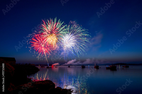 Colorful fireworks explode in a Maine harbor at dusk filled with boats.
