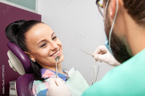 Young beautiful woman patient with white straight teeth sitting in the dental chair and smilling while male doctor examining her teeth with help of dental mirror. Healthcare, medicine.